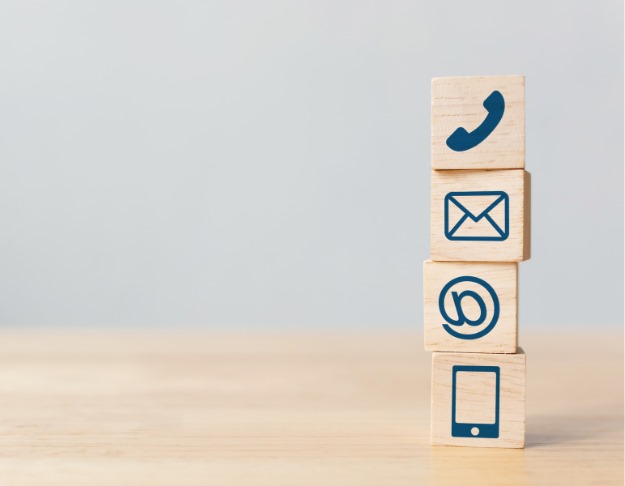 Four wooden blocks with blue icons are stacked vertically. From top to bottom, the blocks display a telephone handset, an envelope, an at symbol, and a smartphone. The background is a light, neutral shade. For more on laboratory equipment costs like those for PCR machines and thermocyclers, contact us today.