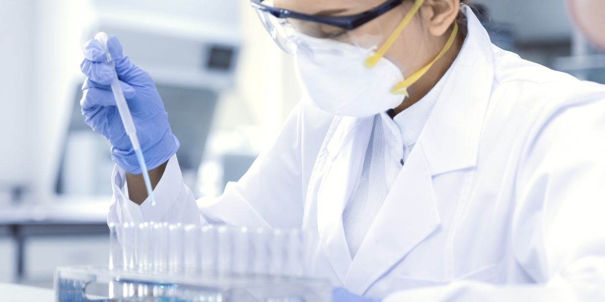 A scientist wearing a white lab coat, safety glasses, a face mask, and blue gloves uses a pipette to transfer liquid into a row of test tubes in a laboratory setting. The lab background is blurred, focusing on the scientist's careful work amidst various laboratory equipments costing thousands.